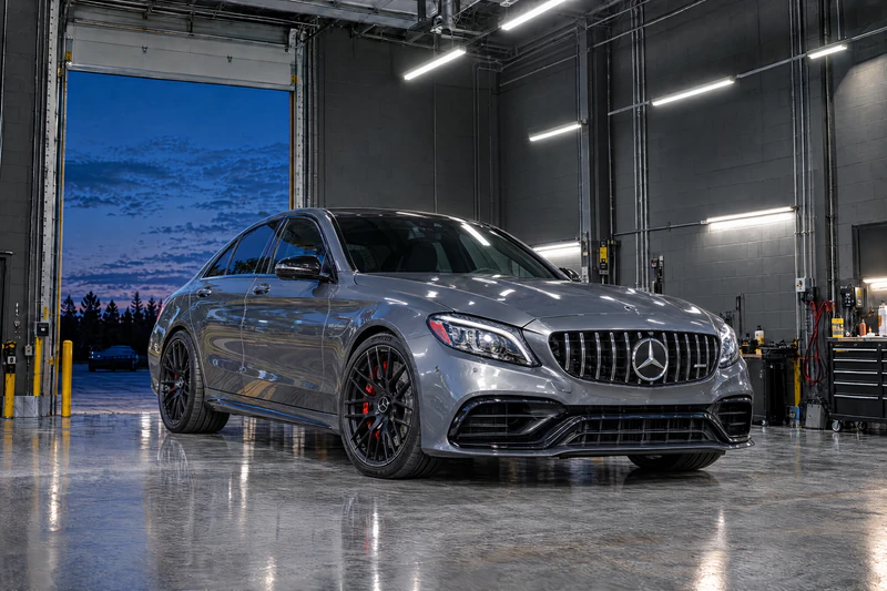 A grey Mercedes-AMG C63 S sedan parked in a clean service bay, roll-up door open at dusk